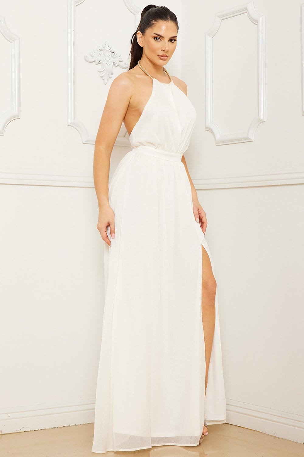 White halter-neck maxi dress with gold neckline detail, backless design, and side slit for formal occasions.