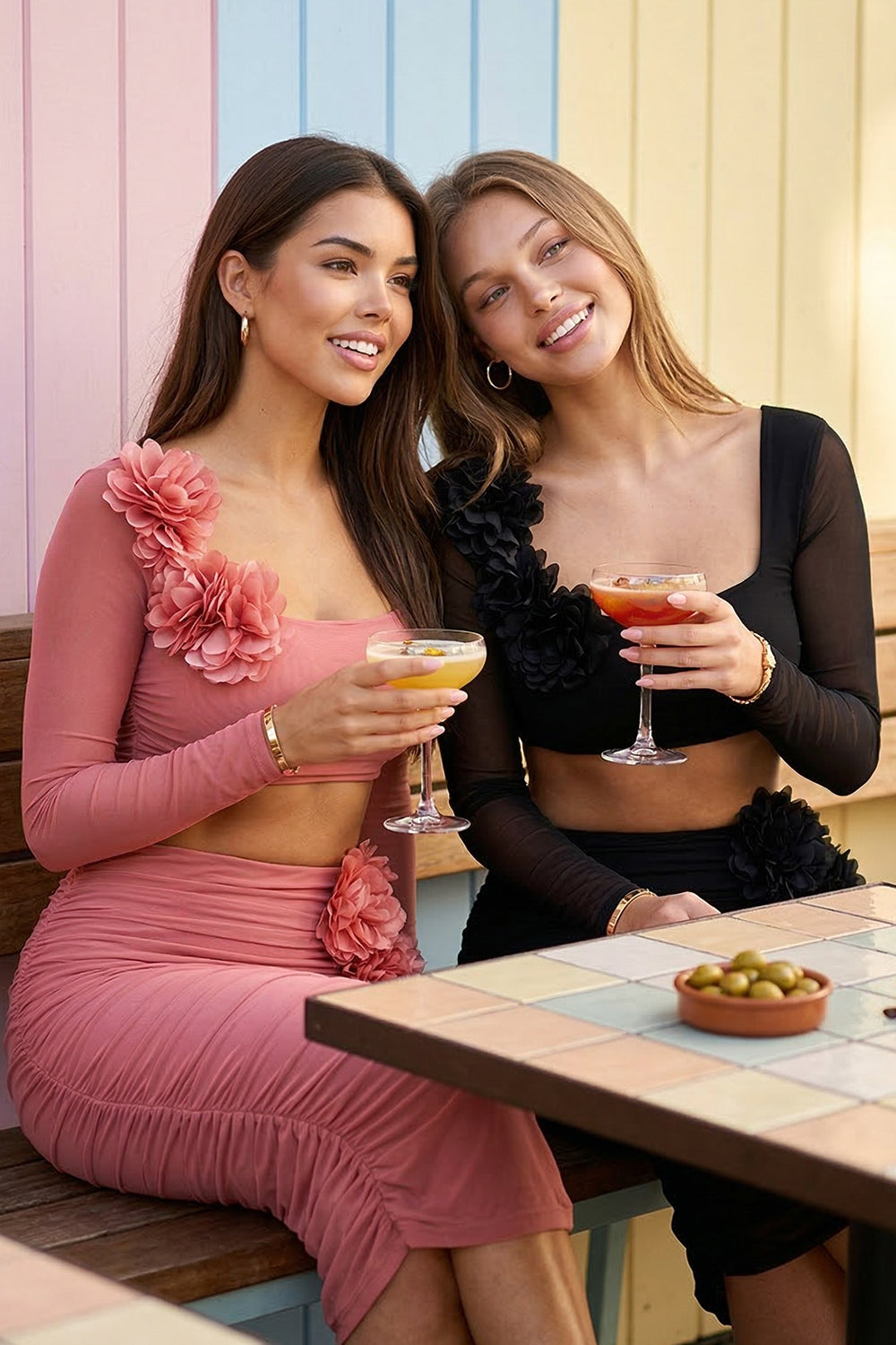 Two women wearing pink and black ruched mesh long-sleeve crop top and midi skirt sets with floral shoulder accents.