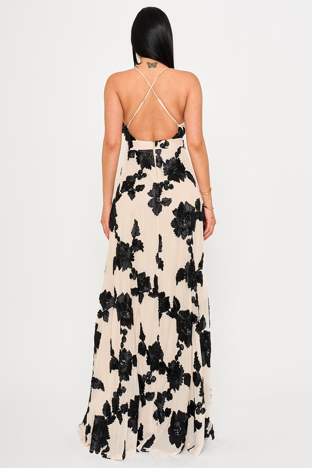 Back view of a beige maxi dress with black floral velvet applique, open back, and crisscross straps by Boutinie.