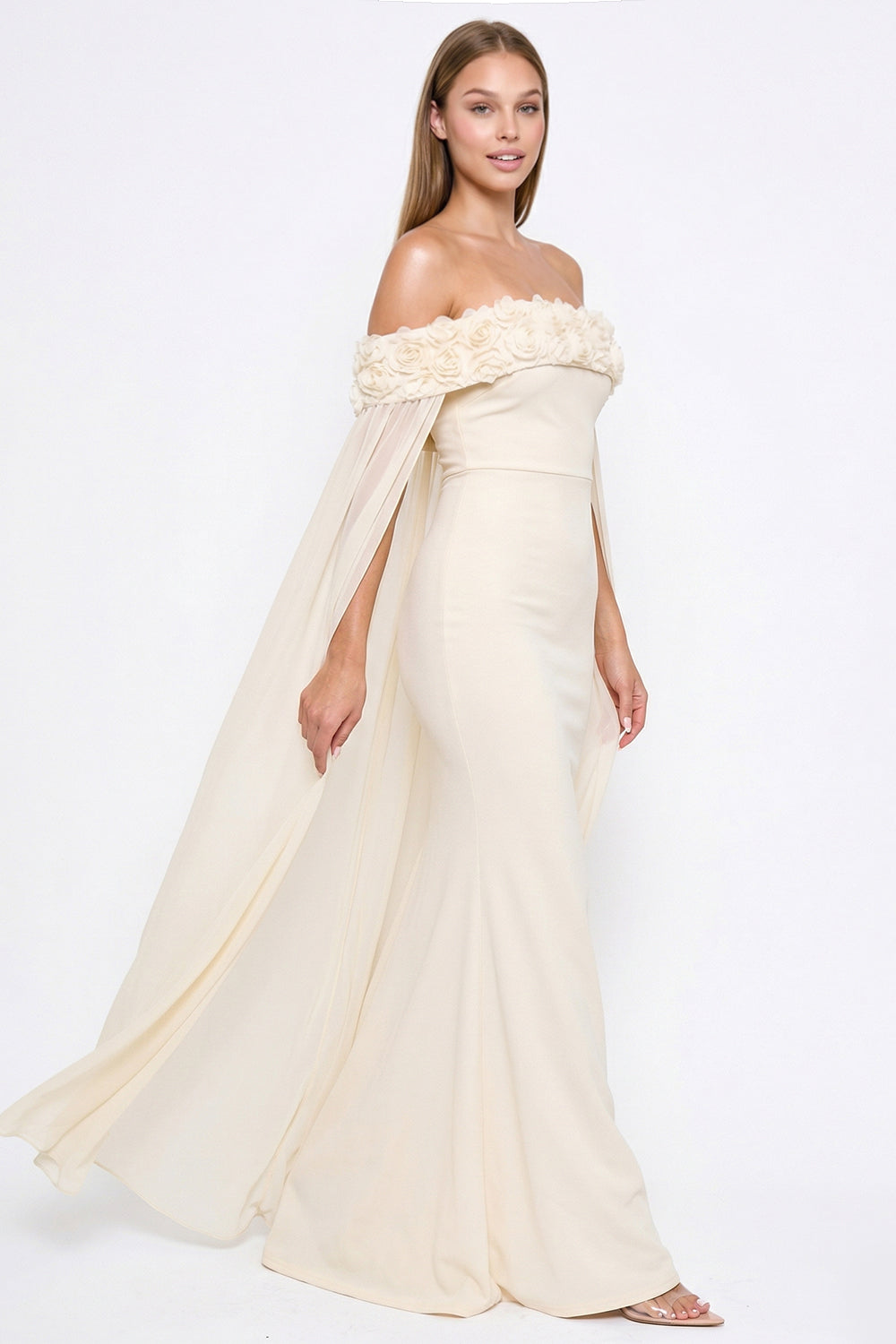 Cream off-shoulder evening gown with floral neckline, flowing cape sleeves, and a sleek floor-length mermaid silhouette.